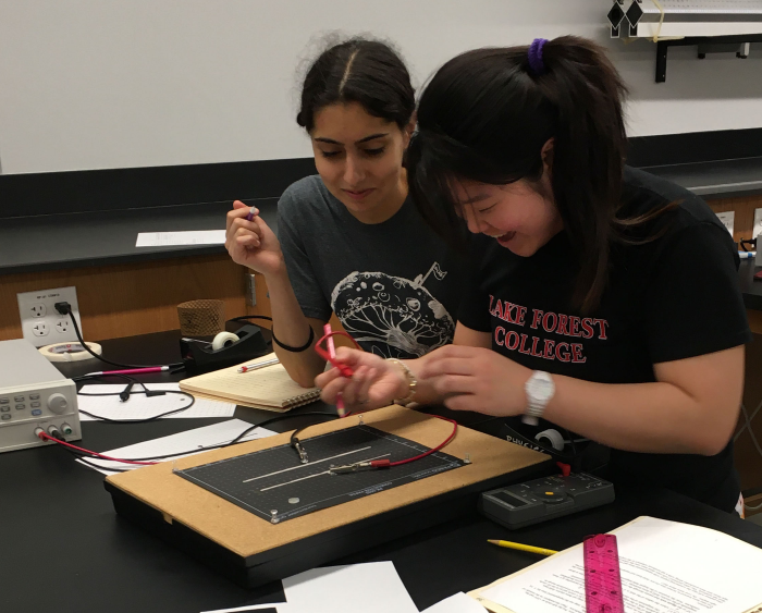 Two female physics students work on a lab project together. they are smiling.